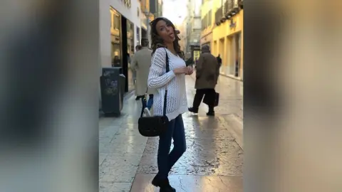 MontseFerrera stands on a city sidewalk wearing a white long-sleeved knit top and blue jeans, sticking her tongue out at the camera.