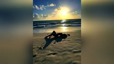 JessycaPage lies naked on the beach at sunset with the ocean waves behind her.