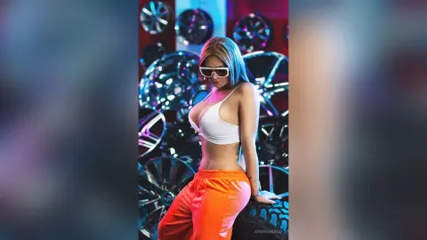 AndreaKalo poses in a white bikini top and orange pants, wearing white sunglasses against a background of shiny chrome car rims.