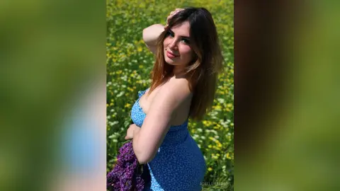 Smiling young woman in a blue floral sundress holding purple flowers in a sunny wildflower field.