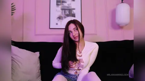 Young woman with long dark hair in a white lace top sitting on a black sofa in a pink aesthetic room.