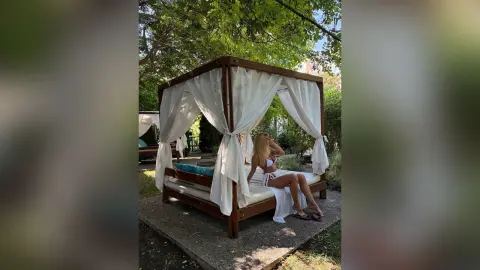 AmaFox — Relaxed woman in white bikini relaxing on a luxury outdoor canopy daybed in a lush green garden setting.