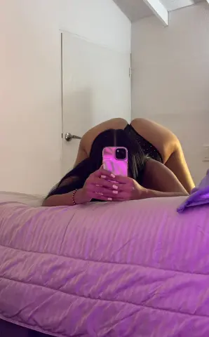 Woman in black lingerie taking a mirror selfie on a bed in a cozy, dimly lit bedroom.