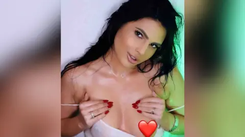 Aalyahh, with long dark hair, pulls at her white lace bra to expose her wet, shiny breasts while looking at the camera.