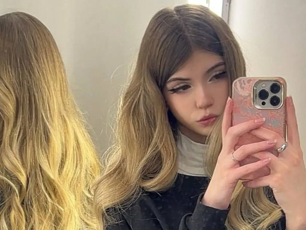 Young woman with long blonde hair taking a mirror selfie in a black turtleneck sweater.