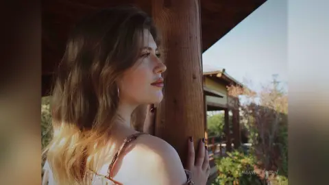 Young woman in a summer dress looking out from a wooden porch during a sunny golden hour afternoon.