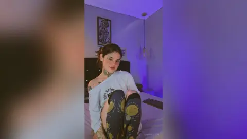 Young woman with tattoos wearing a white t-shirt and patterned leggings in a moody, purple-lit bedroom.