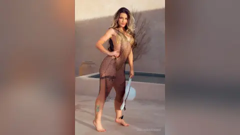 Glamorous woman in a sheer gold mesh maxi dress posing by a luxury poolside at sunset.