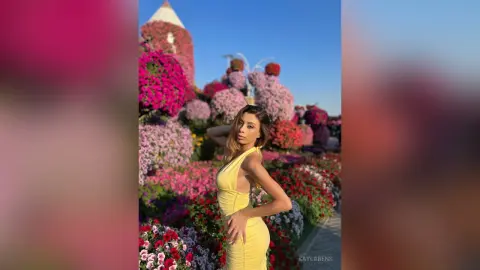 Stylish woman in a yellow summer dress posing amidst vibrant pink and red flower gardens under a clear blue sky.