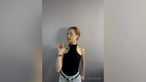 MalliaLucioni poses against a grey wall, wearing a black halter neck top and light blue jeans while holding a pair of glasses near her face.