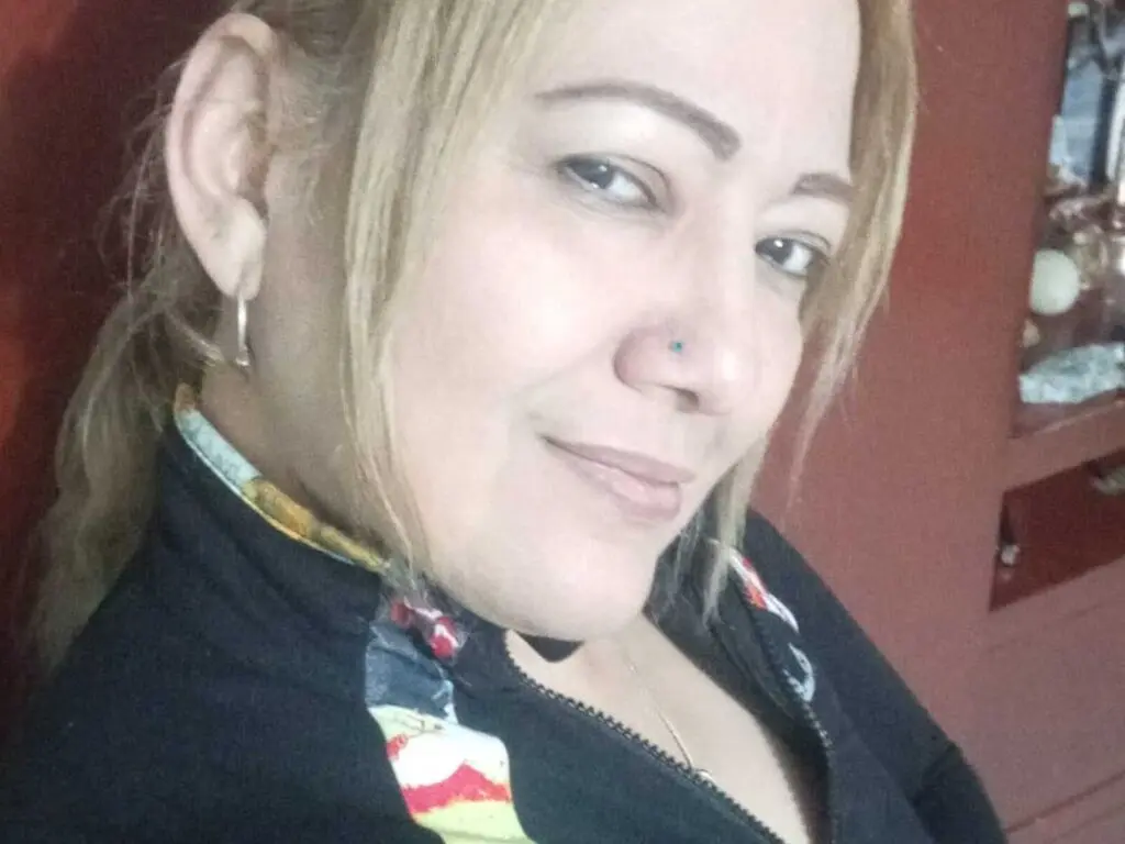 Smiling blonde woman in a black zip-up jacket relaxing in a cozy indoor setting.