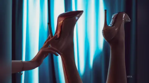 Elegant woman in beige high heels posing against blue curtains in a moody, cinematic setting.