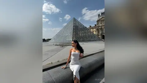 Elegant woman in a white cocktail dress sitting by the Louvre Pyramid in Paris under a blue sky.