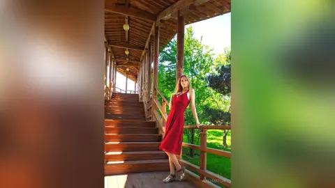Elegant woman in a red dress standing on a wooden staircase in a lush green garden setting.