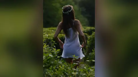 Dreamy woman in a white summer dress and flower crown walking through a lush green field at sunset.