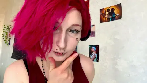 Woman with bright pink hair and facial piercings posing in a room decorated with anime posters.