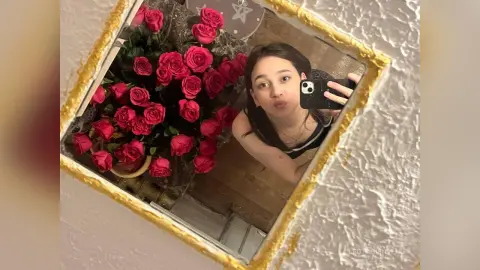 Young woman in a black tank top taking a mirror selfie next to a bouquet of red roses in a cozy room.