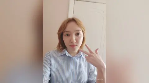 Smiling young woman in a blue striped button-down shirt posing indoors with a peace sign.