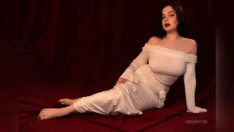 Elegant woman in a white off-the-shoulder long sleeve dress posing against a moody red velvet backdrop.