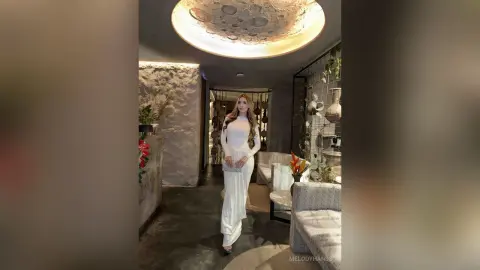 Elegant woman in a white long-sleeve maxi dress standing in a modern, luxury lounge with moody lighting.