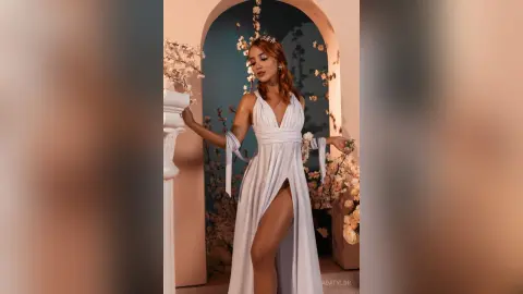 AdaTylor poses in a white slit dress with long sleeves against a blue archway and white flowers.
