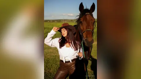 Stylish woman in white blouse and brown cowboy hat posing with a brown horse in a sunny green field.