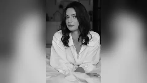 Portrait of a young woman in a white button-down shirt sitting on a bed in a moody black and white aesthetic.