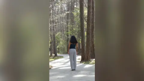 Woman in black crop top and grey trousers walking through a serene, sunlit forest path.