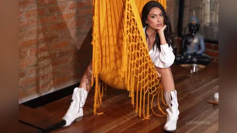 LucyMartin sits on a bright yellow macrame hammock wearing a white blouse and white cowboy boots.