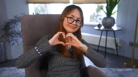 Smiling young woman in a grey sweater making a heart gesture with her hands while sitting in a cozy living room.