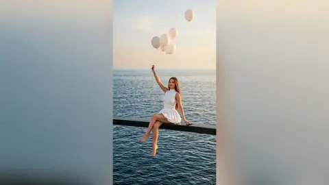 Dreamy woman in a white dress sitting on a pier holding white balloons over the ocean at sunset.