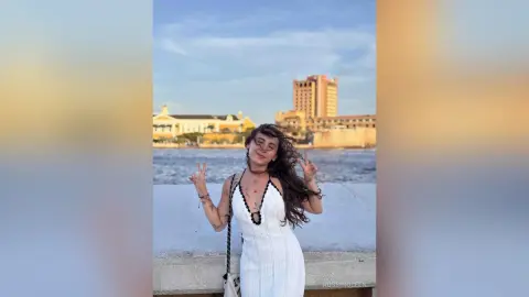 Young woman in a white sundress making peace signs by the ocean near a coastal city at sunset.
