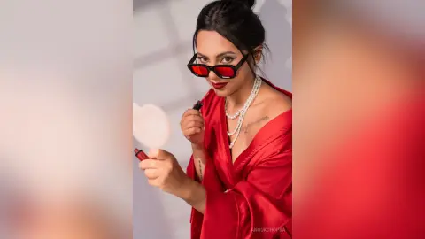 Stylish woman in a red satin robe and red sunglasses posing glamorously against a neutral studio background.