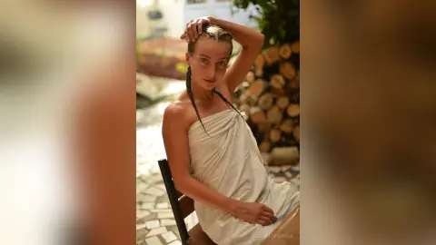 Serene woman in a white draped linen dress sitting outdoors near rustic wood logs.