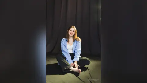 Smiling young woman in a blue striped shirt and black pants sitting on a floor against a dark curtain backdrop.