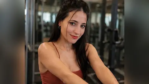 Smiling brunette woman in a rust tank top posing in a bright modern gym setting.