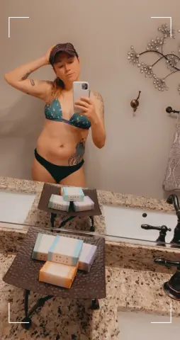 Woman in teal bikini and baseball cap taking a mirror selfie in a modern bathroom setting.