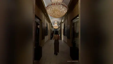 Elegant woman in a backless gown walking through a luxury hallway under glowing crystal chandeliers.
