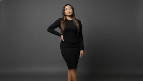 XinemaBonet poses with one hand on her hip, wearing a tight black long-sleeved dress against a dark grey background.