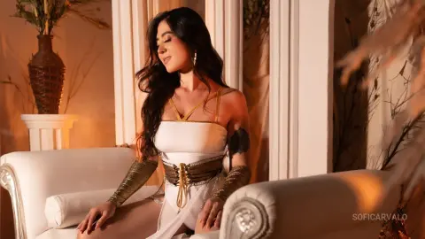 SofiCarvalo sits on a white sofa wearing a white and gold strap outfit with long dark wavy hair.
