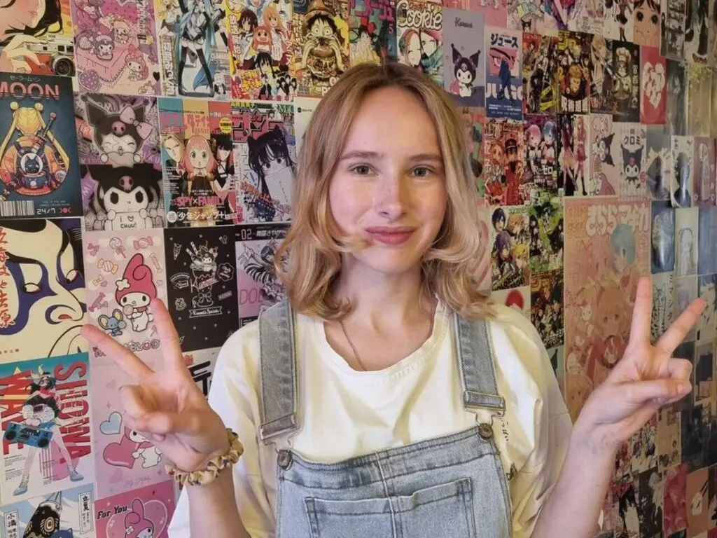 Smiling young woman in white t-shirt and denim overalls posing with peace signs against a colorful anime poster wall.