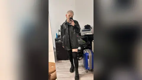 JoyceTreier poses in a mirror wearing a black leather jacket, black shorts, and tall black socks with boots.