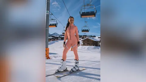 Woman in pink ski jumpsuit standing on a snowy mountain slope near a ski lift under a clear blue sky.