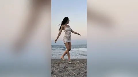 SaraTray stands on a beach wearing a tight, short white patterned sundress, looking off to the side with her long dark hair down.
