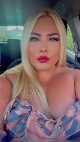 Blonde woman in a pink patterned sundress sitting in a car with a soft, glamorous makeup look.