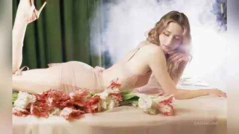 SashaLune lies on a bed wearing a light pink bra and matching panties, surrounded by red and white tulips.