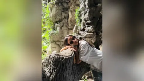 Young woman in a white shirt and sunglasses relaxing against a large rock in a lush green forest setting.