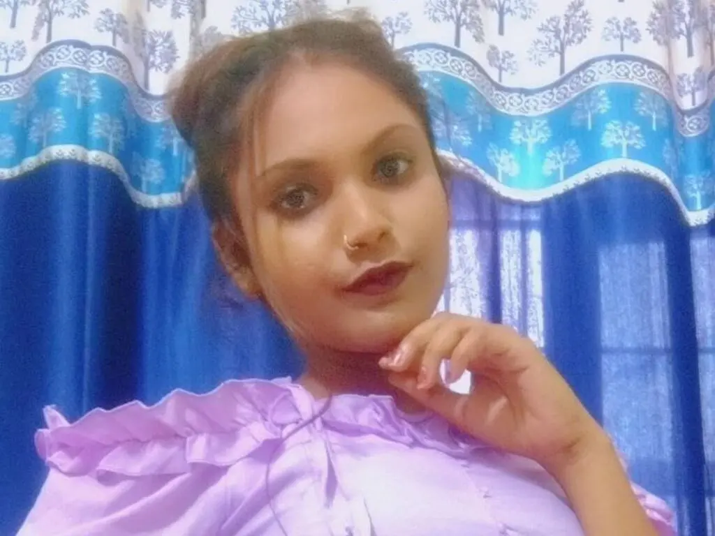 Young woman in a lavender ruffled blouse posing thoughtfully against blue patterned curtains.