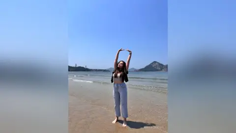 Young woman in a beige crop top and light blue jeans posing with hands in a heart shape on a sunny beach.