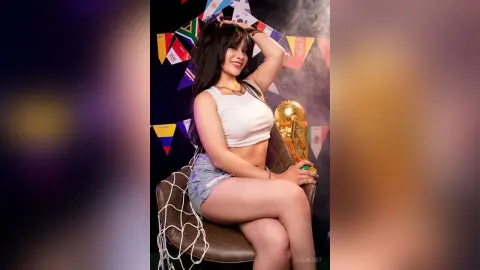 GalaLust poses in a white crop top and denim shorts, sitting on a chair with a gold trophy next to her.